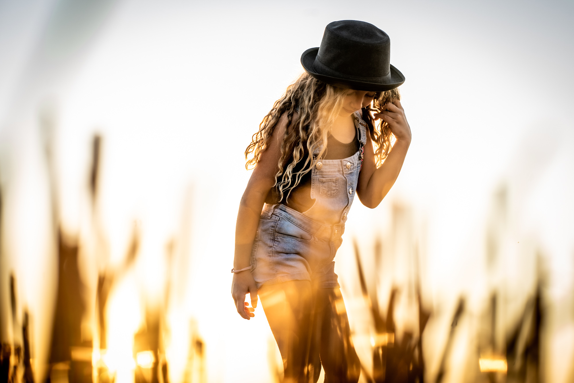 Ein persönliches Portrait mit individuellem Ausdruck. Junge Frau mit schwarzem Hut steht seitlich im weichen Licht, lange blonde Locken fallen über ihre Schulter.
