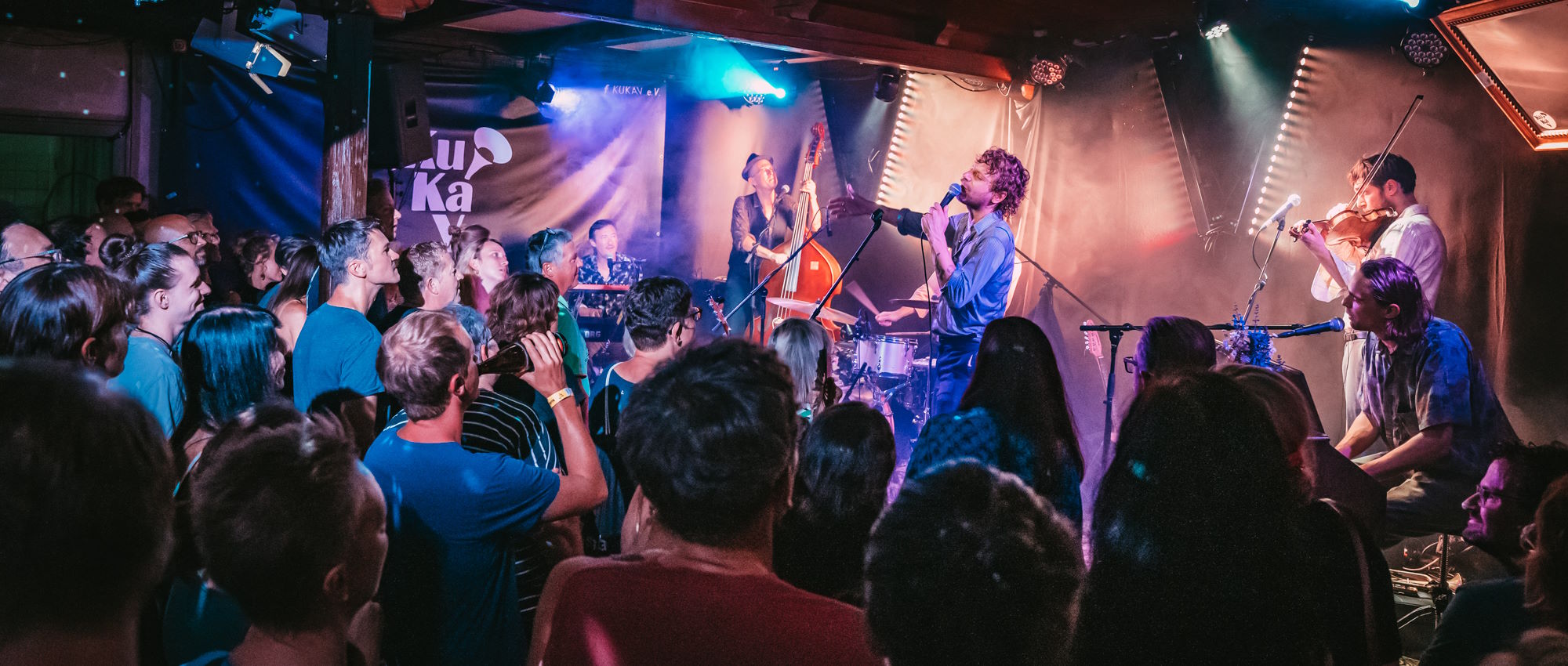 Liveband Voodoo Juergens mit Sänger, Kontrabass und Geige spielt vor dicht gedrängtem Publikum im blau-violetten Bühnenlicht.