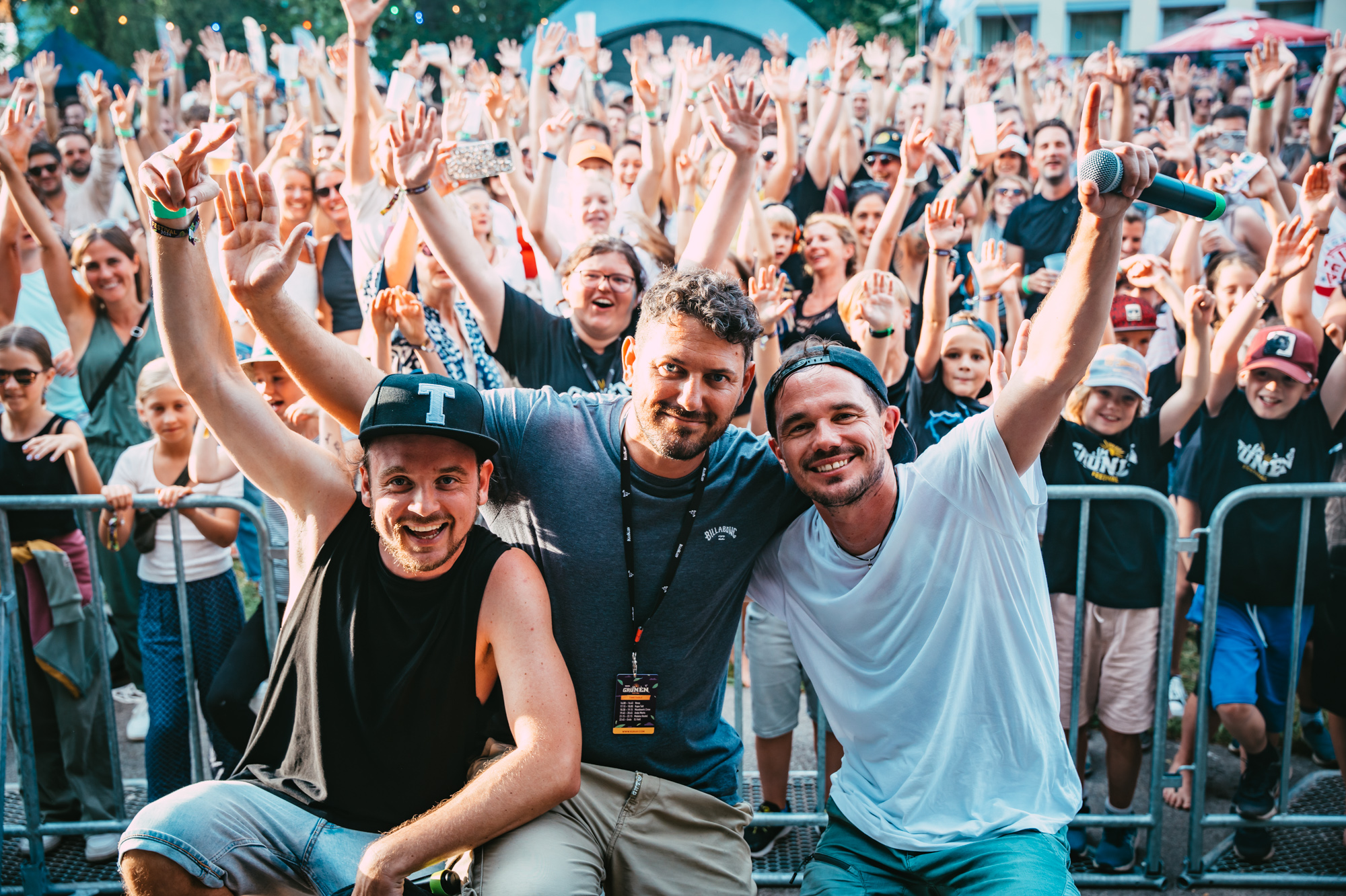 Drei Musiker von der Band Mundwerk Crew jubeln mit erhobenen Armen vor einer großen, ausgelassenen Menschenmenge.